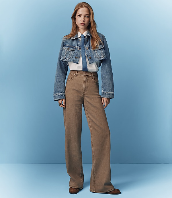 A young woman in a denim jacket and wide-leg pants stands against a solid blue background.