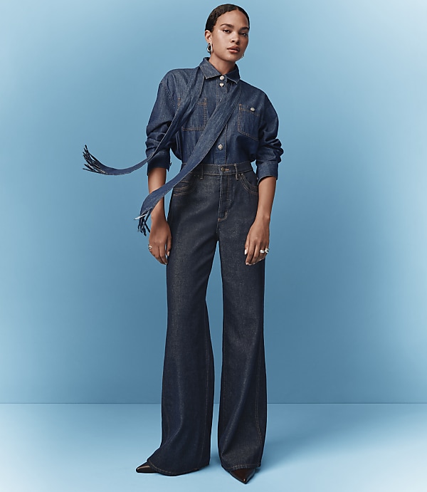 A woman in a denim shirt and wide-leg jeans stands against a solid blue background.