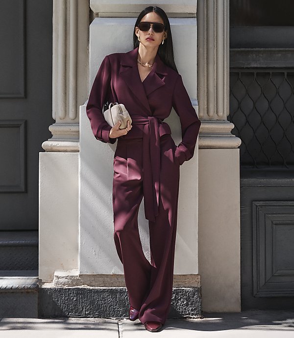 A woman in a burgundy jumpsuit stands against a building with a column and a dark door.