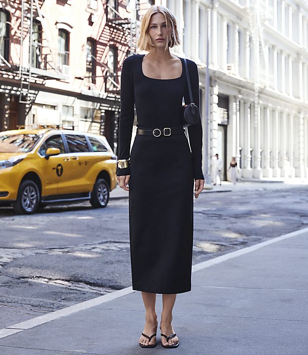 A woman in a black dress stands on a city street with a yellow taxi and buildings in the background.