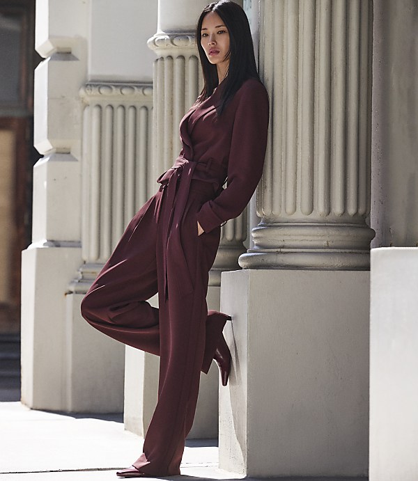 A woman in a maroon jumpsuit leans against a fluted column in front of a building.