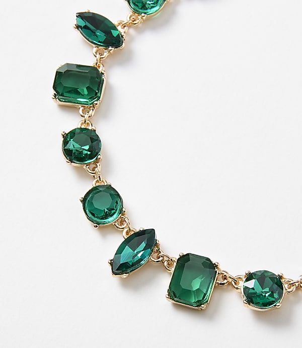 A gold necklace with various emerald-cut and round green gemstones is displayed against a plain white background.