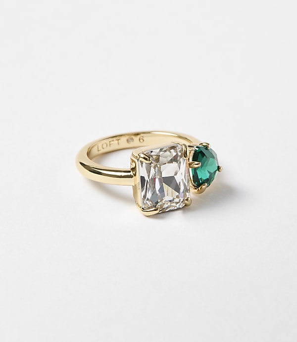 A gold ring with a rectangular clear stone and a heart-shaped green stone sits on a plain white background.