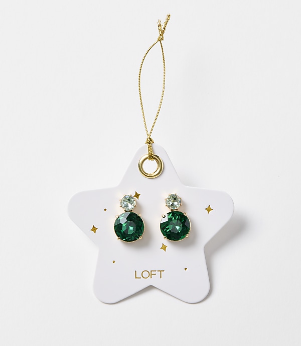 A pair of emerald green gemstone earrings dangle from a gold string attached to a white star-shaped card with gold accents and the "LOFT" logo, set against a plain white background.
