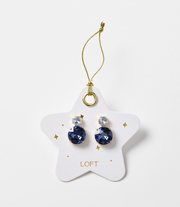 A pair of blue gem earrings are displayed on a white star-shaped card with gold accents and the "LOFT" logo, against a plain white background.