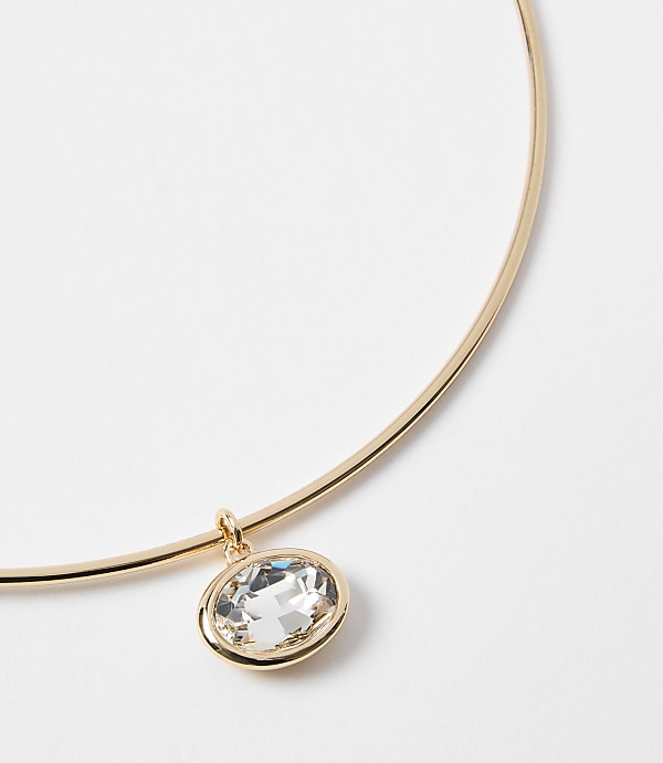 A gold choker necklace with a clear oval pendant hangs against a plain white background.