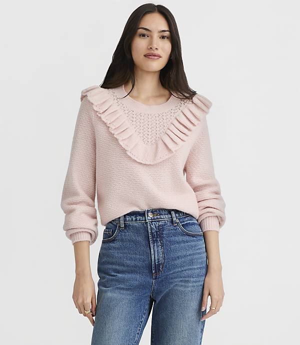 A woman in a pink ruffled sweater and blue jeans stands against a plain white background.