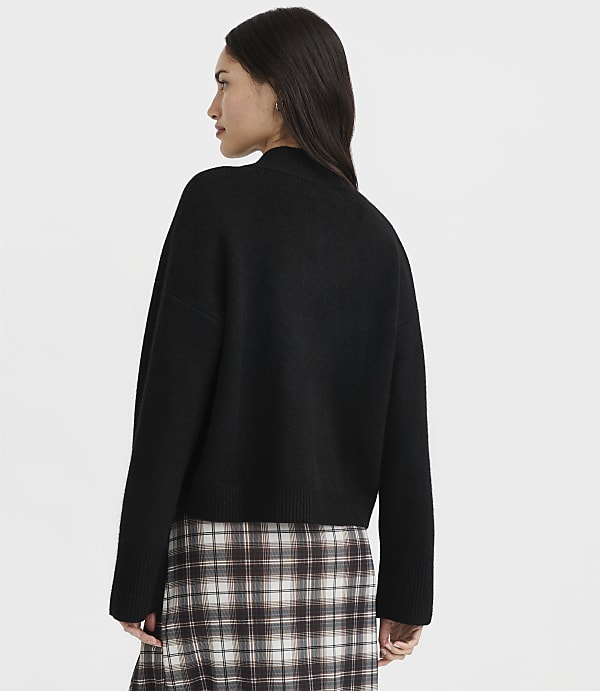 A woman in a black sweater and plaid skirt stands against a plain white background.