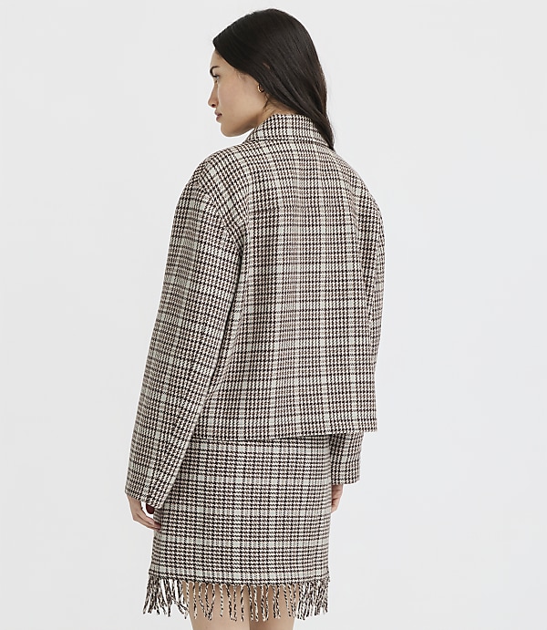 A woman in a plaid jacket and skirt with fringe stands against a plain white background.