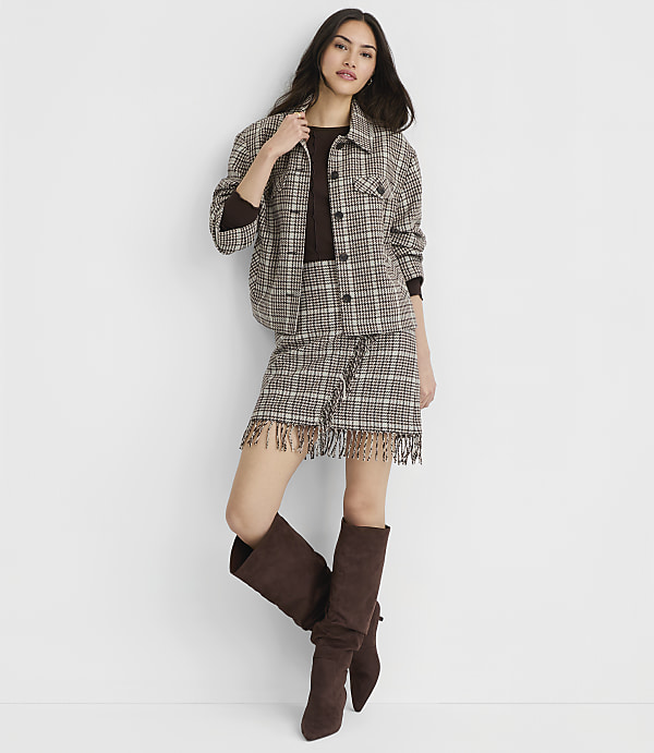 A woman in a plaid jacket and skirt with fringed hem stands against a plain white background, wearing brown boots.