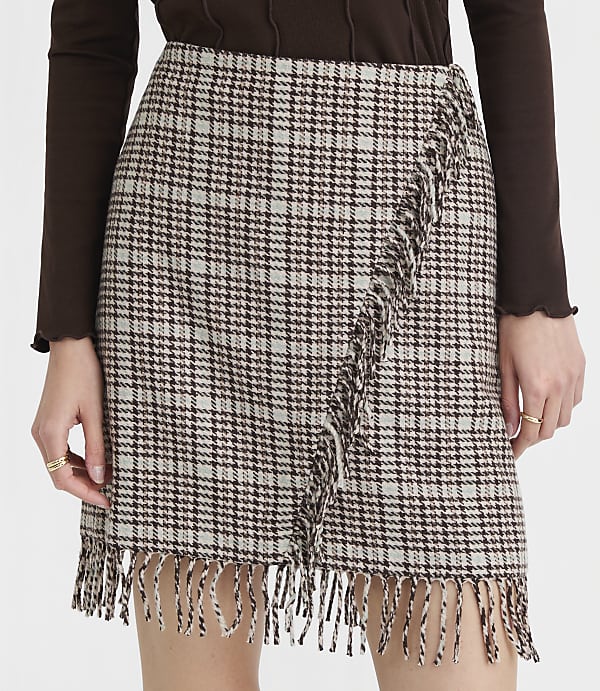 A fringed, plaid mini skirt is worn by a person with rings on their fingers, against a plain brown background.