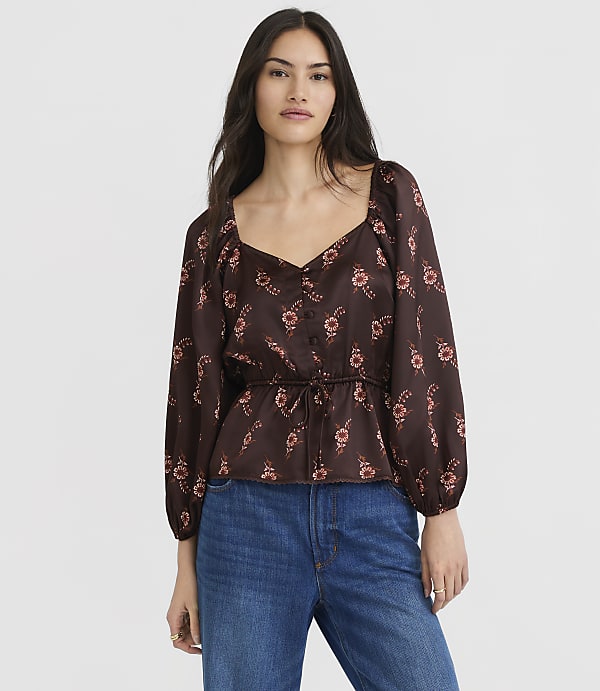 A woman in a floral print blouse and jeans stands against a plain gray background.