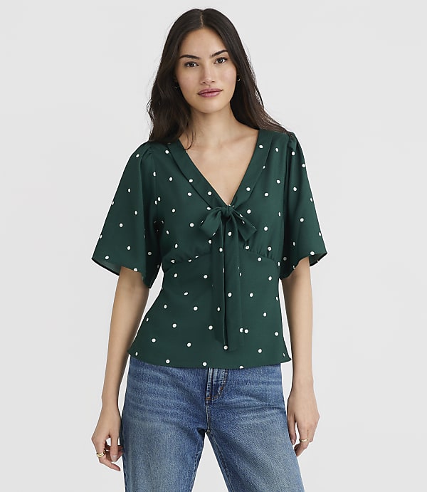 A woman in a green polka dot blouse and blue jeans stands against a plain white background.