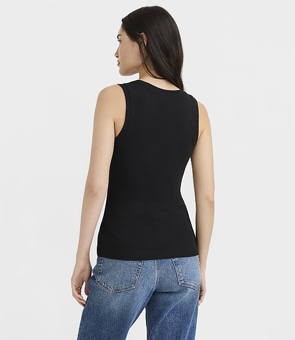 A woman in a black ribbed tank top and blue jeans stands against a plain white background.