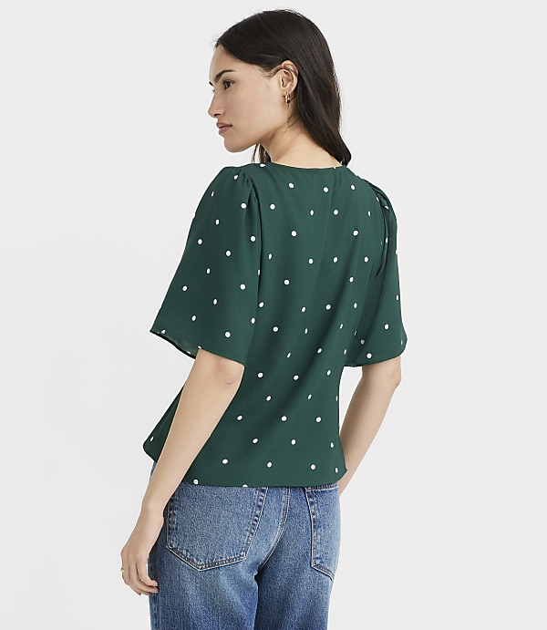 A woman in a green polka dot blouse and blue jeans stands against a plain white background.