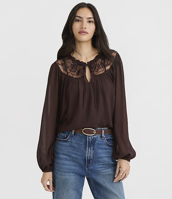 A woman in a brown lace-trimmed blouse and blue jeans stands against a plain white background.