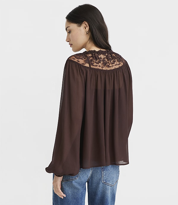 A woman in a brown lace-trimmed blouse and blue jeans stands against a plain white background.