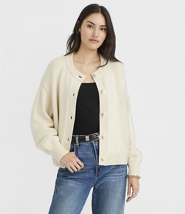 A woman in a cream cardigan and blue jeans stands against a plain white background.