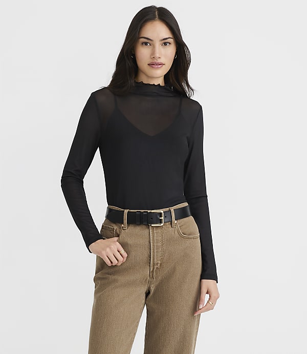 A woman in a black sheer top and brown pants stands against a plain white background.
