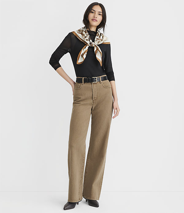 A woman in a black top, brown jeans, and a patterned scarf stands against a plain white background.
