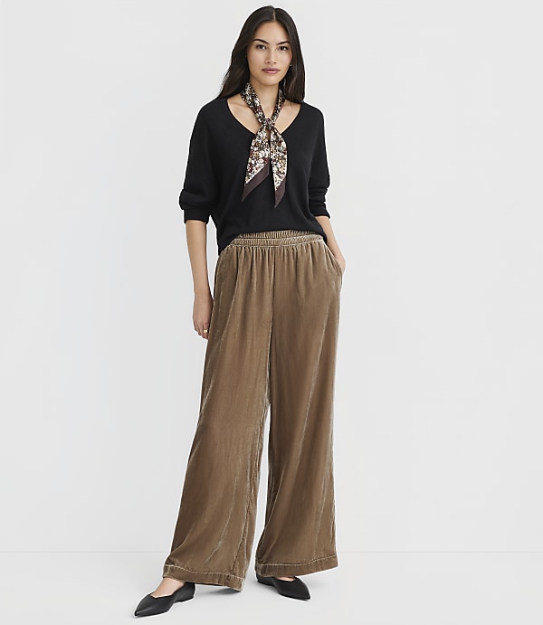 A woman in a black sweater and brown velvet pants stands against a plain white background.