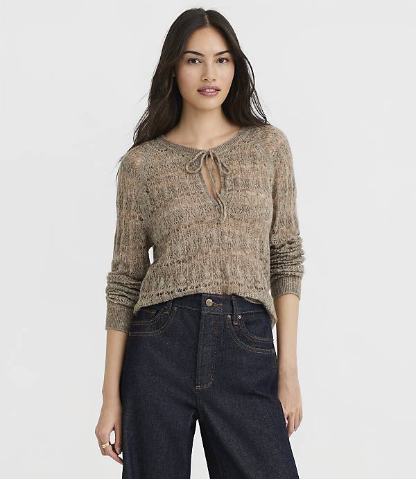 A woman in a light brown, open-knit sweater with a tie at the neck and dark wash jeans stands against a plain white background.