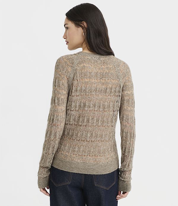 A woman in a sheer, textured brown sweater and dark jeans stands against a plain, light gray background.