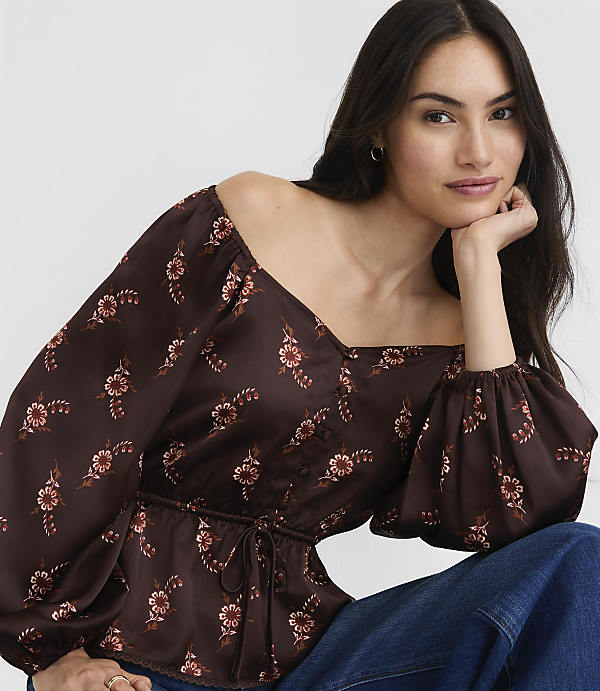 A woman in a floral off-the-shoulder top and jeans rests her chin on her hand against a plain background.