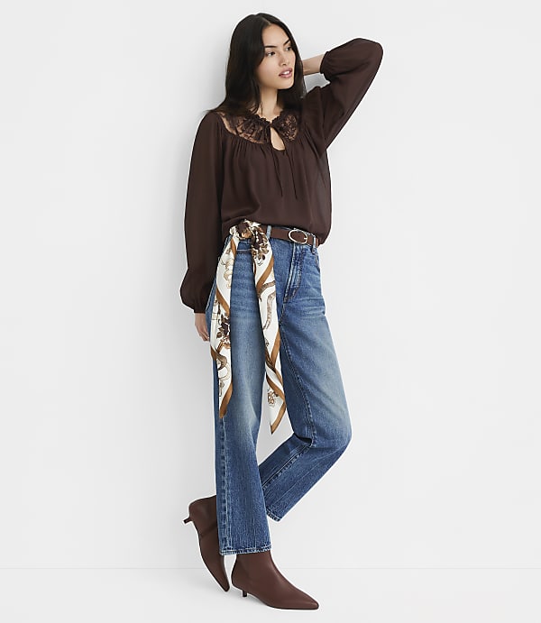 A woman in a brown lace-trimmed blouse and jeans stands against a plain white background.