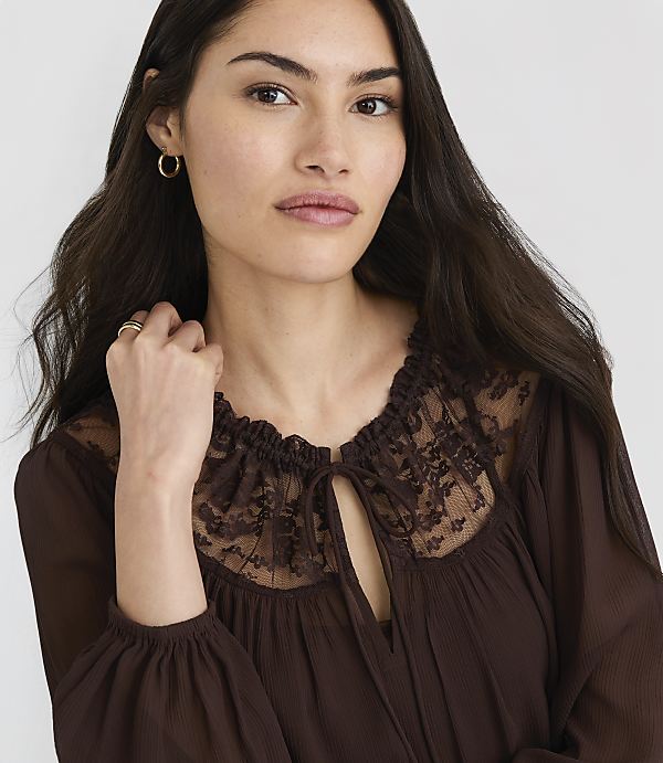 A woman with long dark hair and delicate features wears a brown lace-trimmed blouse against a plain gray background.