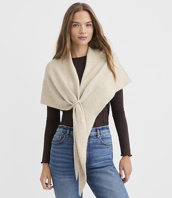 A woman in a brown long-sleeved shirt and blue jeans wears a beige shawl tied at her waist against a plain white background.