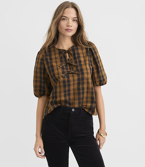 A woman in a brown plaid top with puffed sleeves and black pants stands against a plain gray background.