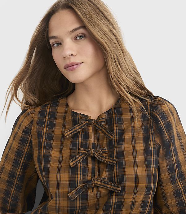 A woman with long, light brown hair wears a brown and black plaid blouse with three bows at the neckline, set against a plain gray background.