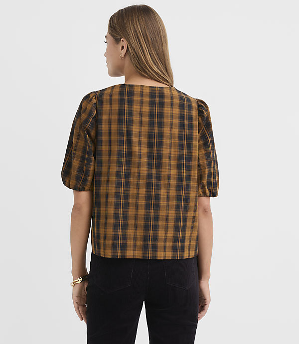 A woman in a brown and black plaid puff-sleeve top stands against a plain white background.