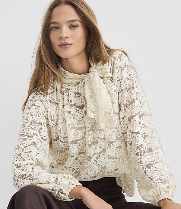 A woman with long brown hair wears a cream-colored lace blouse with a pussy-bow tie, against a plain white background.