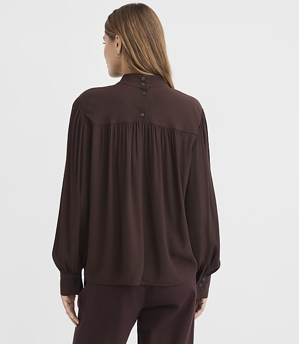 A woman in a brown blouse and pants stands against a plain white background.