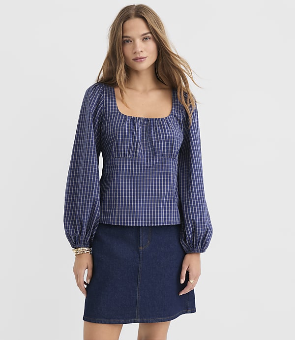 A woman in a blue checkered puff-sleeve top and denim skirt stands against a plain white background.