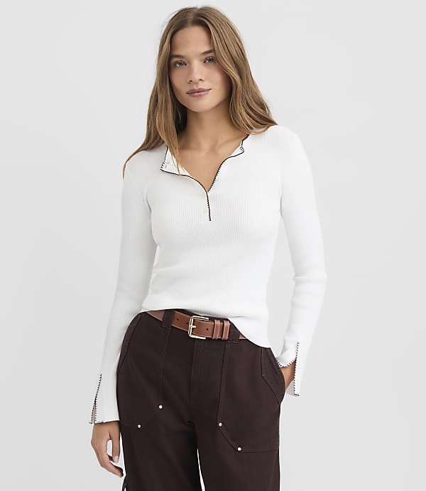 A woman in a white ribbed top and brown pants stands against a plain white background.