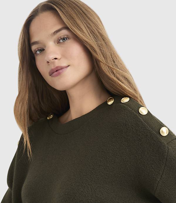 A woman with long, light brown hair wears a dark green sweater with gold buttons on the shoulders, against a plain gray background.
