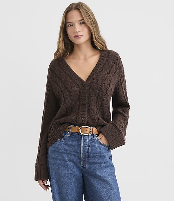 A woman in a brown cable-knit cardigan and blue jeans stands against a plain white background.