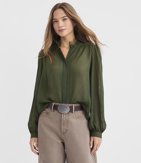 A woman in a green blouse and brown pants stands against a plain gray background.