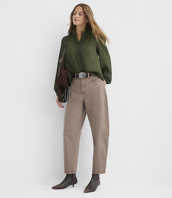 A woman in a green blouse and brown pants stands against a plain white background, accessorized with a brown belt, boots, and a fringed bag.