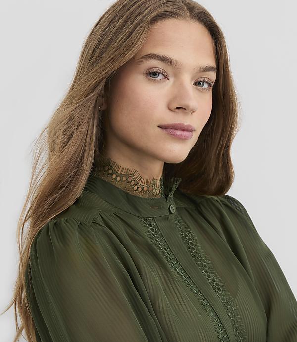 A young woman with long, brown hair and blue eyes looks forward, wearing a green, high-necked blouse with lace trim, against a plain gray background.