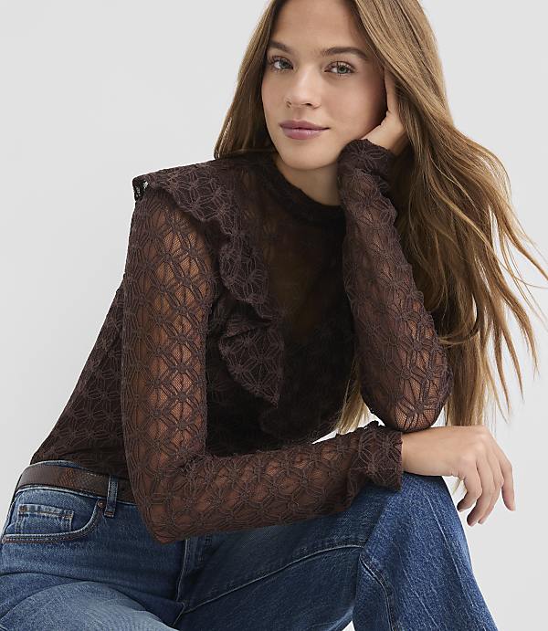 A woman in a brown lace top and blue jeans poses against a plain white background.