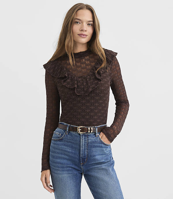A woman in a brown ruffled top and blue jeans stands against a textured white background.