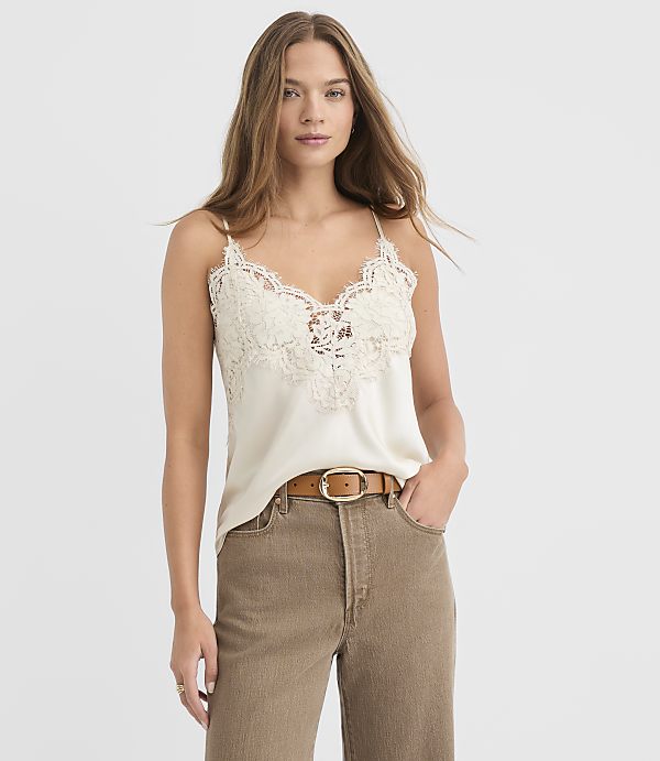A woman in a cream lace camisole and brown pants stands against a plain white background.