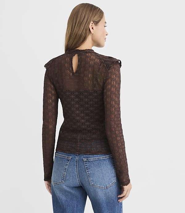 A woman in a sheer, patterned brown top and blue jeans stands against a plain white background.