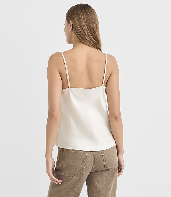 A woman in a cream camisole and brown pants stands against a plain white background.