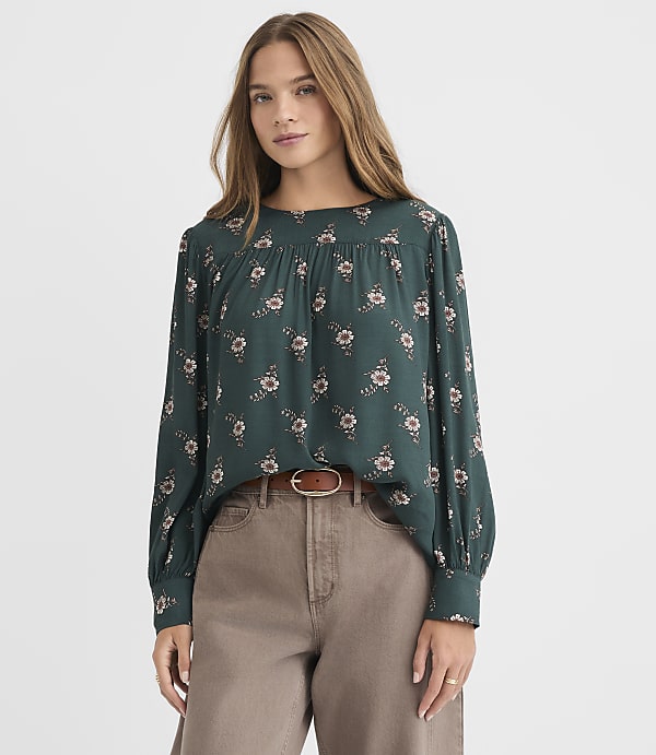 A woman in a dark green floral blouse and brown pants stands against a plain white background.