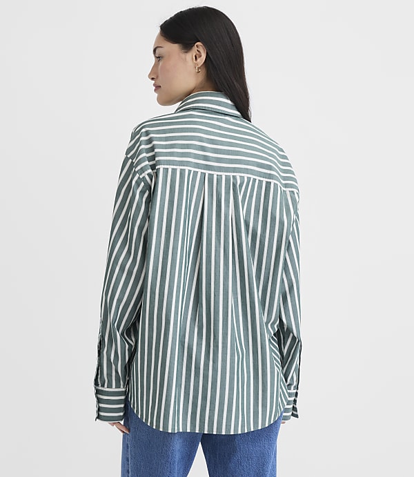 A woman in a green and white striped oversized shirt stands against a plain white background.